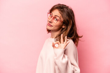 Young caucasian woman isolated on pink background winks an eye and holds an okay gesture with hand. - obrazy, fototapety, plakaty