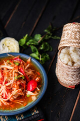 Spicy papaya salad served in a blue dish with sticky rice in a bucket and long beans, dried chilli and vegetables.