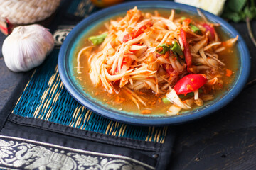 Spicy papaya salad served in a blue dish with sticky rice in a bucket and long beans, dried chilli and vegetables.
