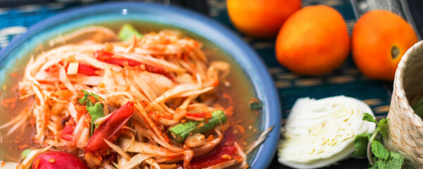 Spicy papaya salad served in a blue dish with sticky rice in a bucket and long beans, dried chilli and vegetables.