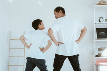 Fototapeta premium Asian young father in white t shirt and his son in white t shirt are showing their backs while looking at each other with silly expression, spending time together at home