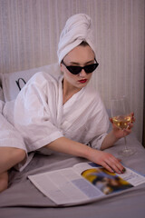 Girl drinking champagne and reading magazine. Attractive woman in bedroom in white bathrobe lies in bed drinking wine and turns over pages of fashion magazine. Close-up view