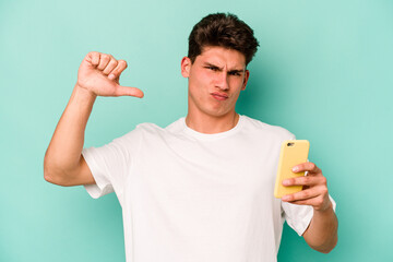 Young caucasian man holding mobile phone isolated on blue background feels proud and self...