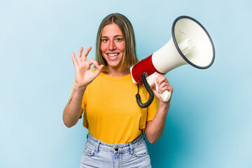 Naklejka premium Young caucasian woman holding a megaphone isolated on blue background cheerful and confident showing ok gesture.