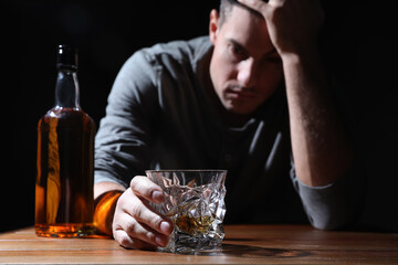 Addicted man at wooden table against black background, focus on glass of alcoholic drink