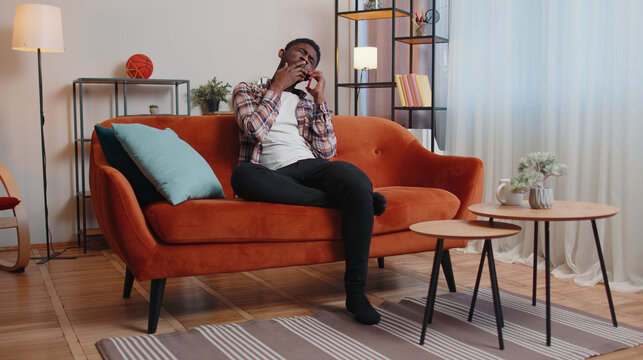 Tired sleepy casual man in shirt talking on mobile phone with friend at home. Sad African American guy freelancer making online conversation speaks on smartphone sitting alone on sofa at living room