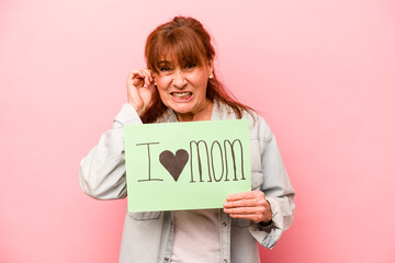 Fototapeta na wymiar Middle age caucasian woman holding I love mom placard isolated on pink background covering ears with hands.