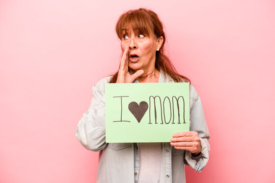 Middle age caucasian woman holding I love mom placard isolated on pink background is saying a secret hot braking news and looking aside