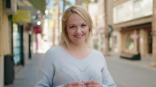 Young blonde woman smiling confident doing heart gesture with hands at street