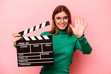 Young caucasian woman holding a clapperboard isolated on pink background smiling cheerful showing...