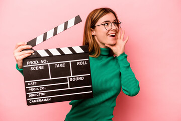 Young caucasian woman holding a clapperboard isolated on pink background shouting and holding palm...