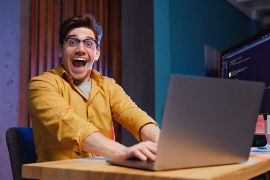 Young surprised impressed winner amazed successful software engineer IT specialist programmer man 20s wearing shirt work at home office writing code on laptop pc computer. Program development concept.