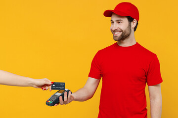 Delivery guy cool employee man in red cap T-shirt uniform work as dealer courier hold wireless bank payment terminal customer process acquire credit card payments isolated on plain yellow background - obrazy, fototapety, plakaty