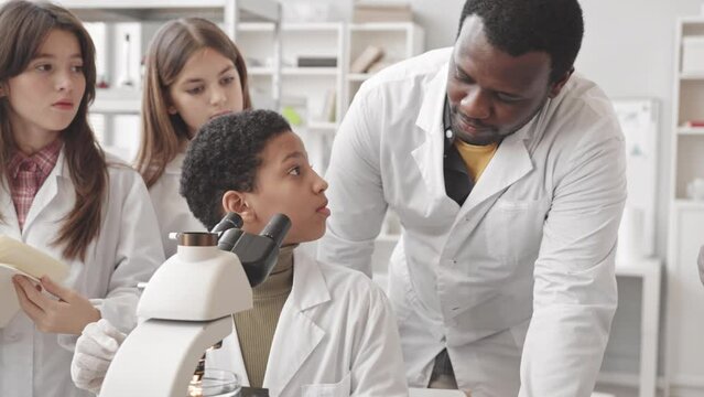 Medium slowmo of African American female pupil in lab coat using microscope at Science class while her classmates and teacher standing nearby watching and taking notes