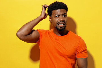 Young confused mistaken puzzled man of African American ethnicity 20s wear orange t-shirt look camera scratch hold head isolated on plain yellow background studio portrait. People lifestyle concept.