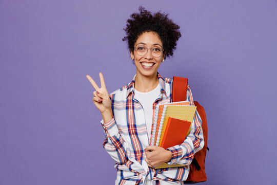 Young girl woman of African American ethnicity teen student in shirt backpack hold books showing victory sign isolated on plain purple background. Education in high school university college concept.