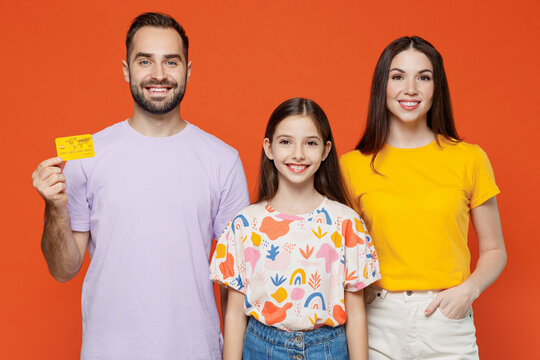 Young Smiling Parents Mom Dad With Child Kid Daughter Teen Girl In Basic T-shirts Hold In Hand Credit Bank Card Isolated On Yellow Background Studio Portrait. Family Day Parenthood Childhood Concept