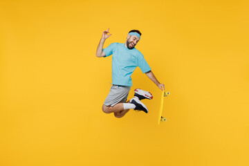 Fototapeta na wymiar Full body side view happy young fitness trainer instructor sporty man sportsman in headband blue t-shirt jump high hold skatebpard isolated on plain yellow background Workout sport motivation concept