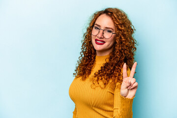 Young ginger caucasian woman isolated on blue background showing victory sign and smiling broadly.