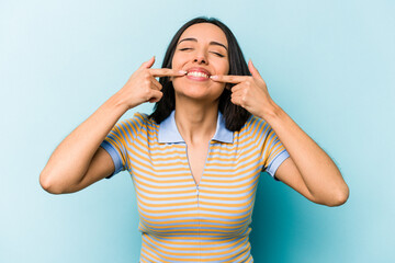 Young hispanic woman isolated on blue background smiles, pointing fingers at mouth.