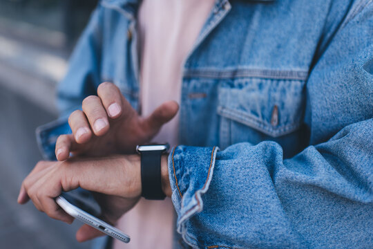Cropped image of millennial user checking meeting time on digital smartwatch waiting friend in travel city, unrecognizable hipster guy in jeans wear holding cellular gadget reading info on wrist clock