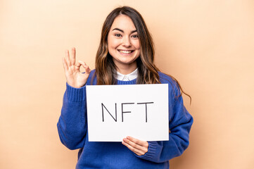 Young caucasian woman holding a NFT placard isolated on beige background cheerful and confident...