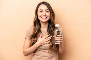 Young caucasian woman holding a bottle of water isolated on beige background laughs out loudly keeping hand on chest.