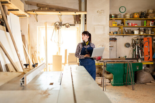 Carpenter woman using laptop with blueprints and planning work in her carpentry workshop