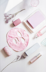 Beauty concept with natural cosmetic products, skin care, pink facial sheet mask and flowers at white background with natural light. Healthy skincare with nourishing products. Top view.