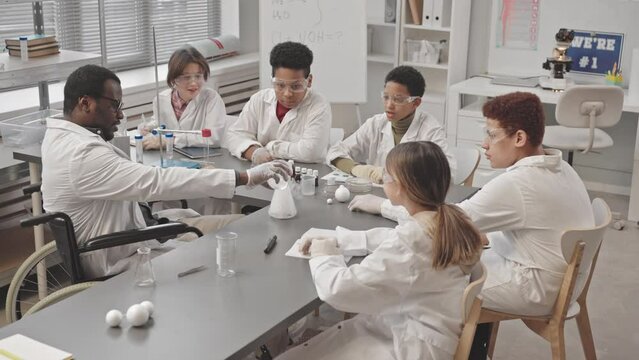 Medium slowmo of African American teacher of Chemistry in wheelchair performing lab experiment to multiethnic group of pupils, adding water to glass flask with dry ice and having smoke as reaction