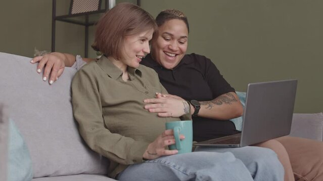 Medium slowmo of happy interracial lesbian couple expecting baby video chatting on laptop with friend showing baby ultrasound image, sitting together on couch at cozy home