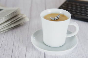 Kaffeebecher in Schrägansicht mit einem Papierschiffchen drin, auf Holzuntergrund, Tastatur und Zeitung im Hintergrund, horizontal