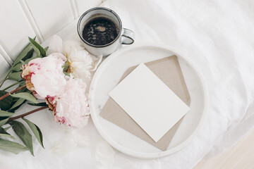 Blooming pink peonies flowers, bouquet lying on table. White wooden wall. Blank greeting card mockup on marble tray. Cup of coffee, breakfast in bed. Wedding or birthday celebration, gift concept.