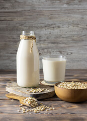 Glass bottle with barley milk on a light brown wooden board. Barley grains in a wooden bowl and a glass of milk in the background. Light wooden background