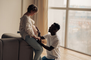 African man kneels before caucasian pregnant woman and touches her belly.