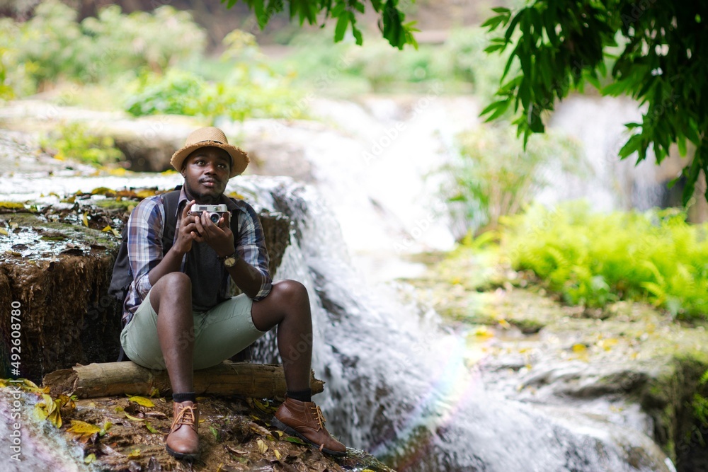 Wall mural african man traveller holding a film camera with backpack sitting and relaxing freedom at the waterf