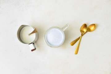 Glass cup of Charcoal Latte, frothing pitcher with milk and spoons on light table