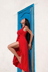 holiday mood. brunette woman in red summer dress stands fashion near white wall background with blue door at sunny day. travel concept, free space