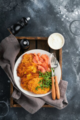 Potato pancakes. Fried homemade potato pancakes or latkes with cream, green onions, microgreens, red salmon and sauce in rustic plate on old black table background. Rustic style. Top view.