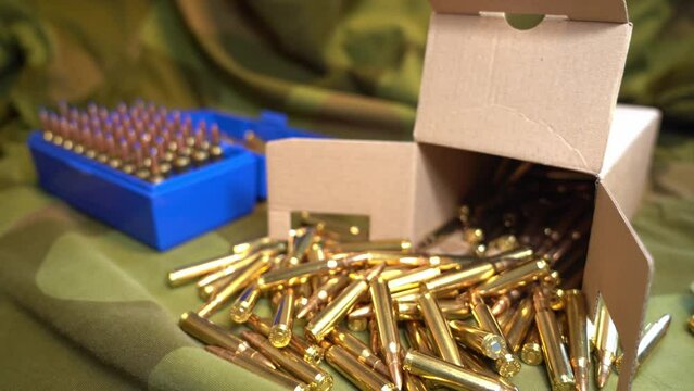Open cardboard box full of 223 military cartridges laying on camouflage jacket and blue ammunition box in background - 3006 cartridges laying on pocket in the end - Slow slider shot left to right