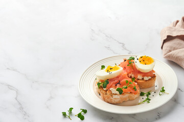 Sandwich with delicious toppings, smoked salmon, eggs, herbs and microgreens radish, black sesame seeds over white plate on white marble table background. Healthy open sandwich superfood. Top view.