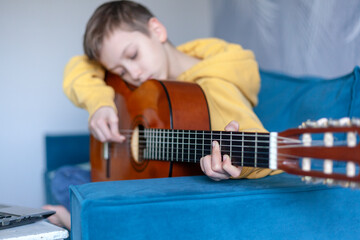 Handsome kid dressed casual jeans and yellow sweatshirt watches video lesson on playing the guitar on the cozy sofa at home.