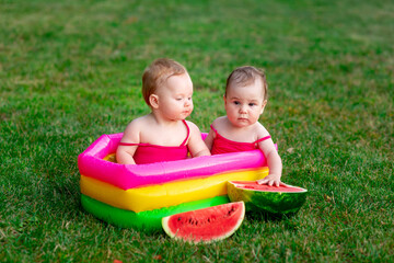 two children of joyful twins swim in an inflatable pool in summer on the green grass with a watermelon, space for text