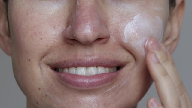 very close up 4 k video of young woman applying white cream on face, on cheek. happy woman is smiling, big front teeth. daily skin routine. natural skin look. beauty and care concept