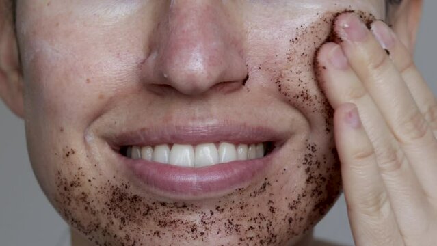 close up video how a woman is applying a man massaging coffee scrub on her face. smiling woman is doing at home skin routine. body and face care. beauty concept. no eyes visible. spa and wellness.