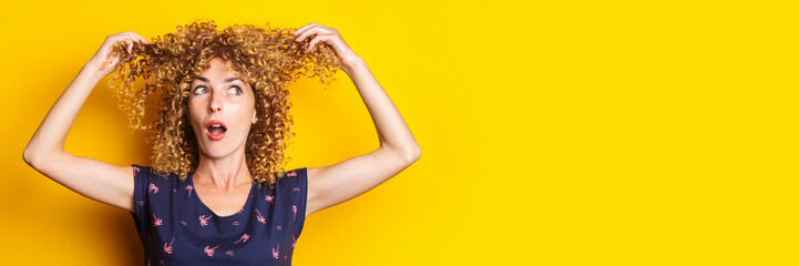 surprised curly young woman lifts her hair up on a yellow background. Banner.
