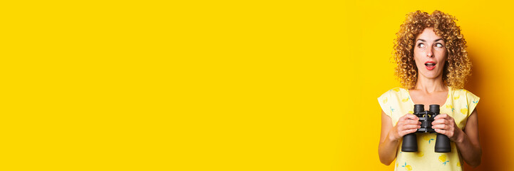 surprised curly young woman with binoculars on yellow background. Banner.