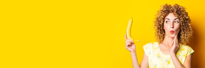 curly young woman in shock looks at a banana on a yellow background. Banner.