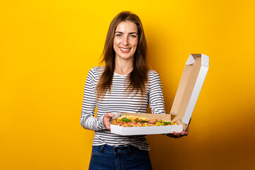 smiling young woman holding package with hot fresh pizza on yellow background