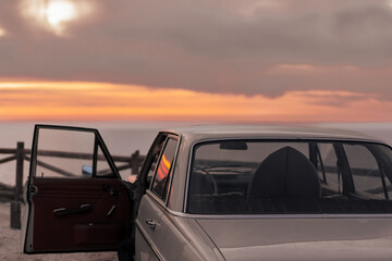 Vintage car parked with a surfboard inside in front of the ocean at the sunset